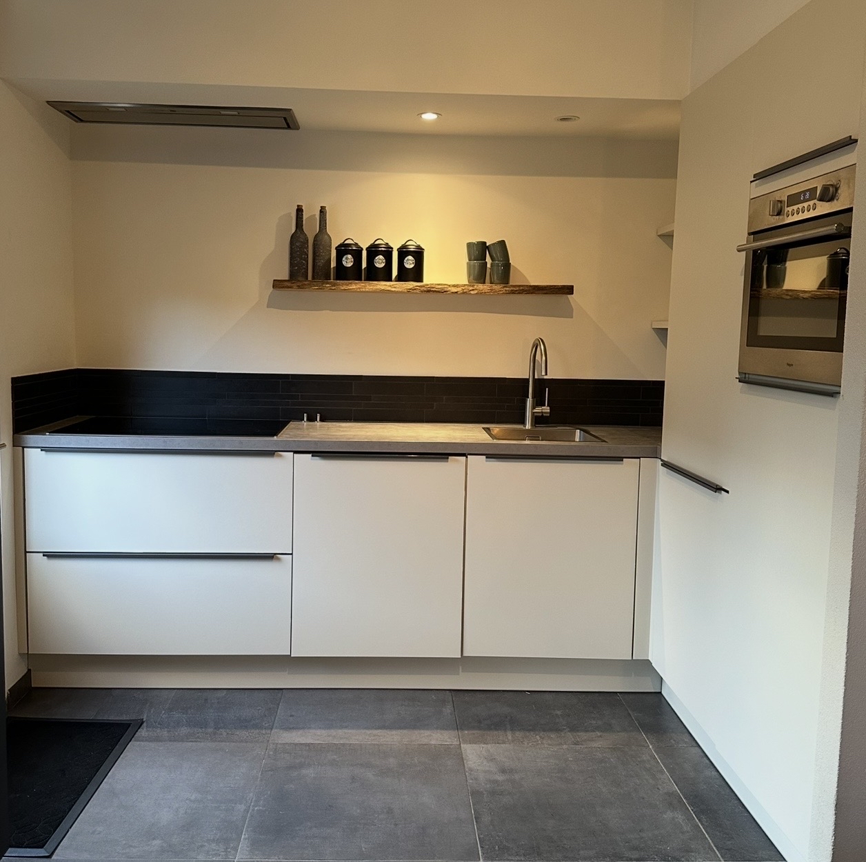 Compacte witte keuken met een donkere achterwand en een houten wandplank