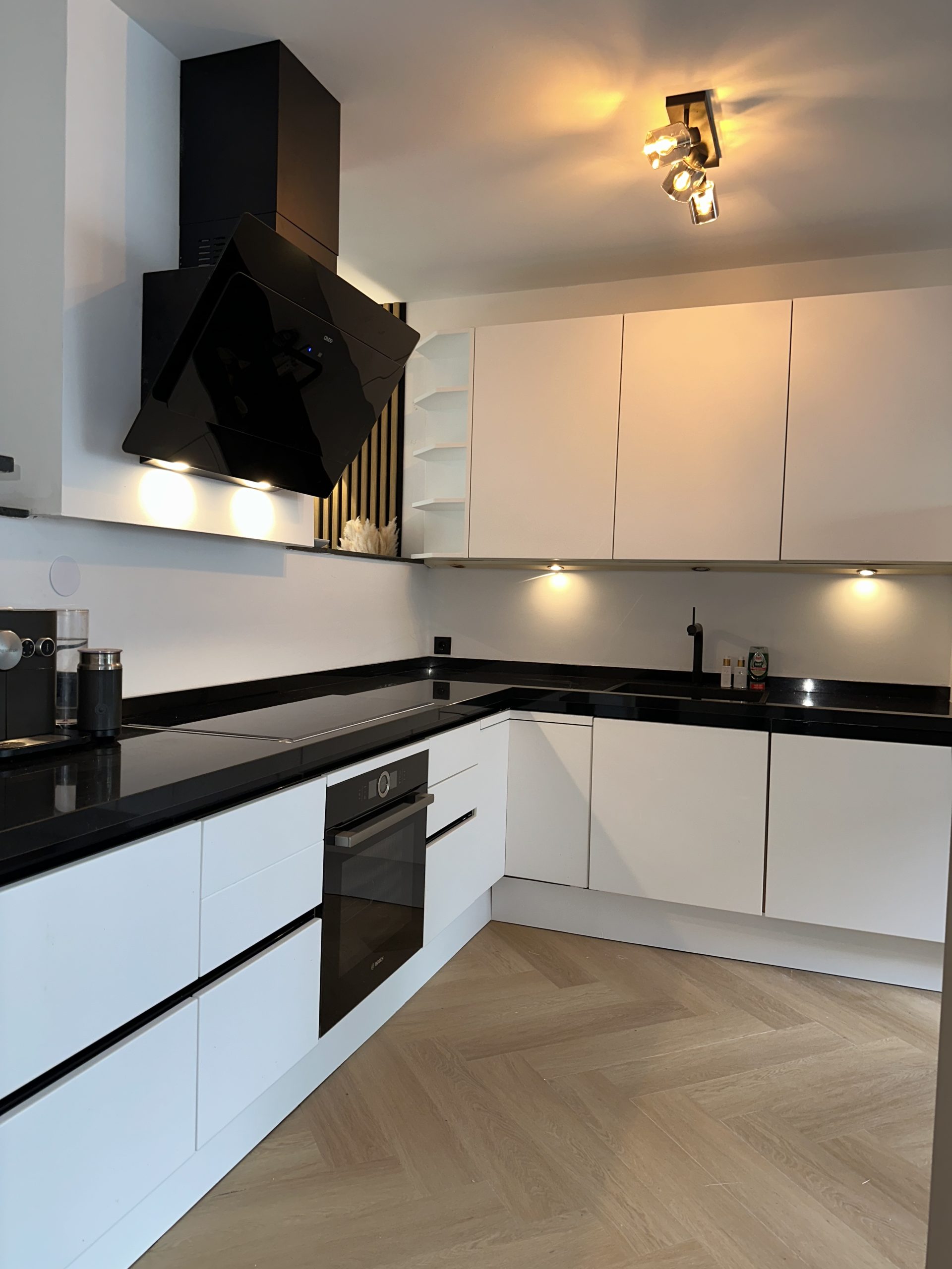 Lichte keuken met witte greeploze fronten, zwart werkblad en houtlook vloer
